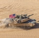 An aerial view of a U.S. Army M1 Abrams main battle tank moving at speed across a desert terrain, kicking up a dust cloud behind it. The tank is flying an American flag from its turret and appears to be in an active operational or training environment. The desert surface shows multiple vehicle track marks indicating prior activity in the area.