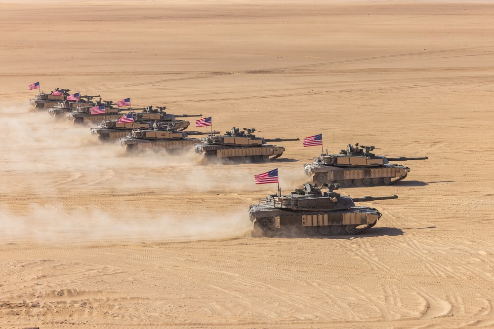 A column of M1 Abrams main battle tanks advances across an open desert terrain in a wedge or echelon formation. Each tank displays a United States flag, and the vehicles are kicking up dust clouds as they move. The image appears to depict a U.S. Army armored unit conducting a desert training exercise or operational movement.