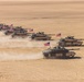 A column of M1 Abrams main battle tanks advances across an open desert terrain in a wedge or echelon formation. Each tank displays a United States flag, and the vehicles are kicking up dust clouds as they move. The image appears to depict a U.S. Army armored unit conducting a desert training exercise or operational movement.
