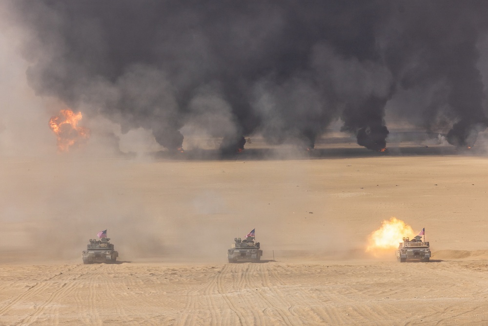 Three American military tanks are advancing across a desert terrain in a line formation, with at least one tank visibly firing its main gun. In the background, multiple large fires produce thick black smoke columns rising into the sky, consistent with burning oil wells or ordnance detonations. Each tank displays a small American flag, indicating U.S. military affiliation.