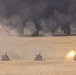Three American military tanks are advancing across a desert terrain in a line formation, with at least one tank visibly firing its main gun. In the background, multiple large fires produce thick black smoke columns rising into the sky, consistent with burning oil wells or ordnance detonations. Each tank displays a small American flag, indicating U.S. military affiliation.