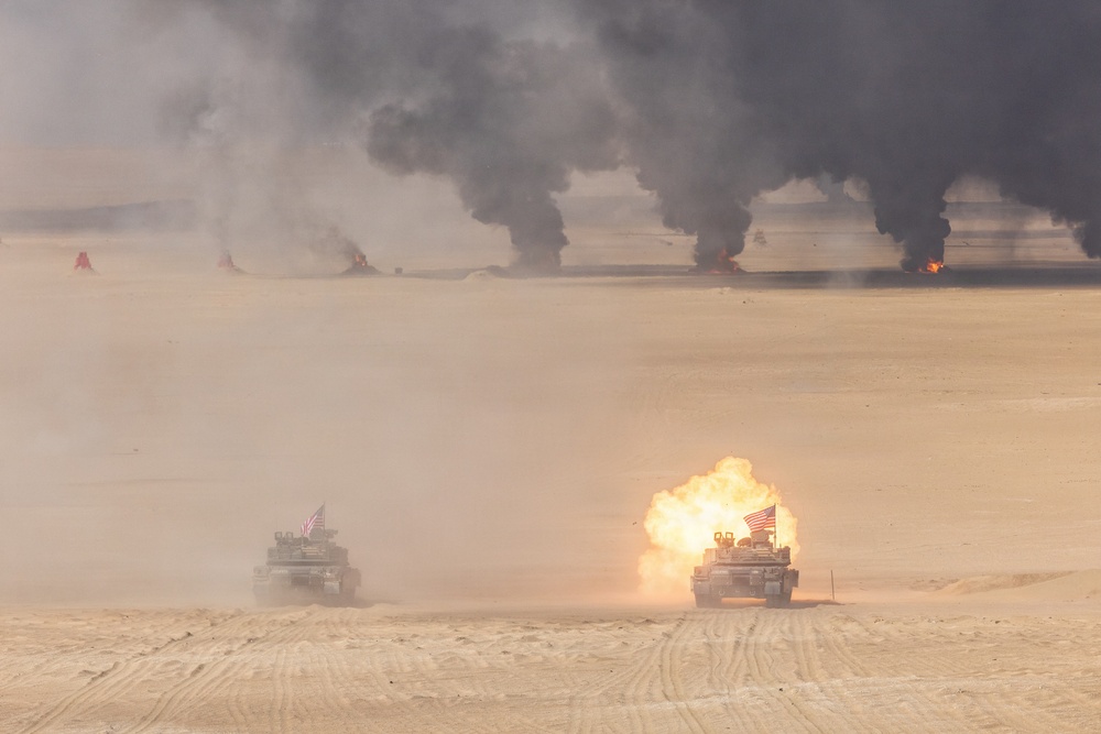 Two military tanks bearing American flags are operating in a desert environment, with one tank visibly firing its main gun producing a large muzzle blast. Multiple fires and columns of black smoke are visible in the background across the flat desert terrain. The scene appears to be a live-fire military exercise or combat operation.