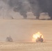 Two military tanks bearing American flags are operating in a desert environment, with one tank visibly firing its main gun producing a large muzzle blast. Multiple fires and columns of black smoke are visible in the background across the flat desert terrain. The scene appears to be a live-fire military exercise or combat operation.