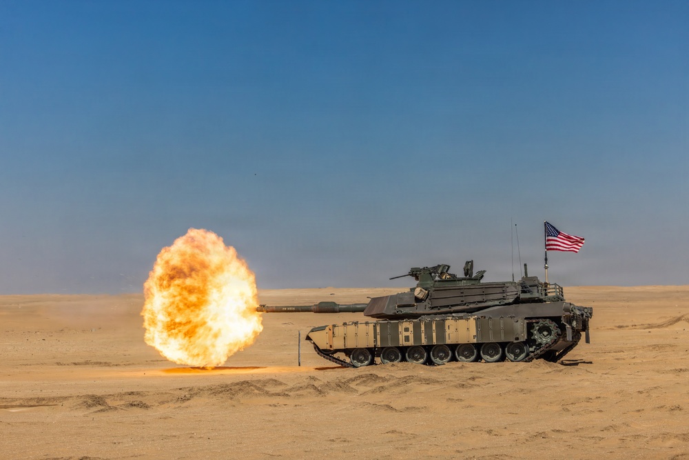 A U.S. Army M1 Abrams main battle tank is firing its main gun in a desert environment, producing a large muzzle blast fireball. An American flag is mounted on the rear of the tank. The scene appears to be a live-fire training exercise or demonstration in an arid, sandy terrain.