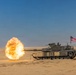 A U.S. Army M1 Abrams main battle tank is firing its main gun in a desert environment, producing a large muzzle blast fireball. An American flag is mounted on the rear of the tank. The scene appears to be a live-fire training exercise or demonstration in an arid, sandy terrain.