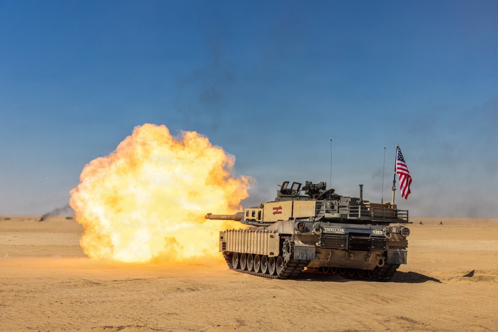 An M1 Abrams main battle tank is firing its main gun in a desert environment, producing a large muzzle blast with orange and yellow flames. The tank displays an American flag on its rear and bears visible unit markings. The image appears to document a live-fire exercise or combat operation in an arid, sandy terrain.