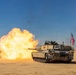 An M1 Abrams main battle tank is firing its main gun in a desert environment, producing a large muzzle blast with orange and yellow flames. The tank displays an American flag on its rear and bears visible unit markings. The image appears to document a live-fire exercise or combat operation in an arid, sandy terrain.