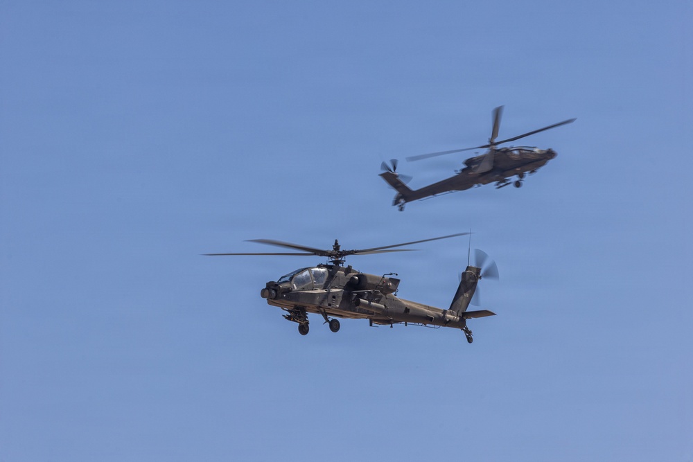 Two military attack helicopters are flying in formation against a clear blue sky. Both aircraft appear to be AH-64 Apache helicopters based on their distinctive tandem cockpit configuration, stub wings, and nose-mounted sensor systems. The foreground helicopter is in sharp focus while the background helicopter shows motion blur, suggesting high-speed flight.