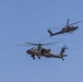 Two military attack helicopters are flying in formation against a clear blue sky. Both aircraft appear to be AH-64 Apache helicopters based on their distinctive tandem cockpit configuration, stub wings, and nose-mounted sensor systems. The foreground helicopter is in sharp focus while the background helicopter shows motion blur, suggesting high-speed flight.