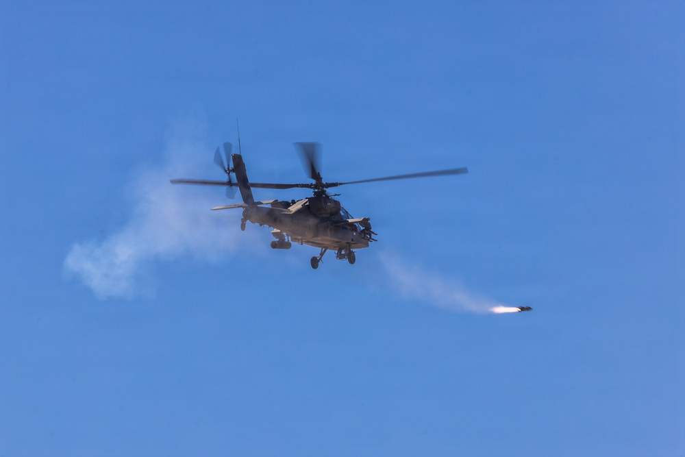A military attack helicopter is captured mid-flight against a clear blue sky, actively firing a rocket or missile. A visible smoke trail extends from the launched projectile toward the right side of the frame. The image appears to document a live-fire exercise or combat operation.