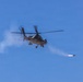 A military attack helicopter is captured mid-flight against a clear blue sky, actively firing a rocket or missile. A visible smoke trail extends from the launched projectile toward the right side of the frame. The image appears to document a live-fire exercise or combat operation.
