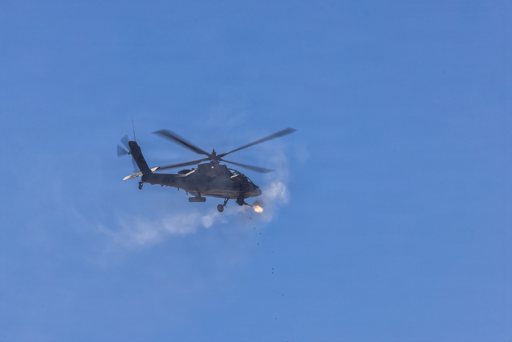 A military attack helicopter is photographed in flight against a clear blue sky. The helicopter appears to be actively firing a weapon, as evidenced by visible muzzle flash and smoke/exhaust cloud near the front of the aircraft. Debris or spent casings are visible falling below the aircraft.