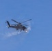 A military attack helicopter is photographed in flight against a clear blue sky. The helicopter appears to be actively firing a weapon, as evidenced by visible muzzle flash and smoke/exhaust cloud near the front of the aircraft. Debris or spent casings are visible falling below the aircraft.