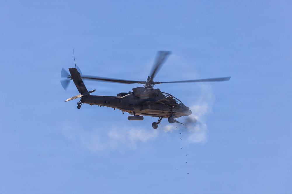 A military attack helicopter, consistent with an AH-64 Apache design, is photographed in flight against a clear blue sky. The helicopter appears to be actively firing its weapons system, as evidenced by a visible smoke plume and debris/projectiles falling below the aircraft. The image is captured from a ground-level perspective looking upward.