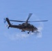 A military attack helicopter, consistent with an AH-64 Apache design, is photographed in flight against a clear blue sky. The helicopter appears to be actively firing its weapons system, as evidenced by a visible smoke plume and debris/projectiles falling below the aircraft. The image is captured from a ground-level perspective looking upward.