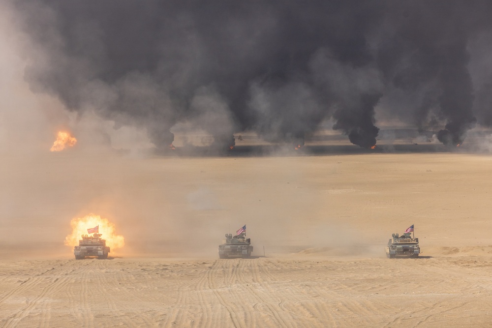 Three military tanks bearing American flags advance across a desert terrain in a line formation. The leftmost tank is firing its main gun, producing a visible muzzle blast. In the background, multiple large fires generate dense black smoke columns across the horizon.