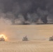 Three military tanks bearing American flags advance across a desert terrain in a line formation. The leftmost tank is firing its main gun, producing a visible muzzle blast. In the background, multiple large fires generate dense black smoke columns across the horizon.