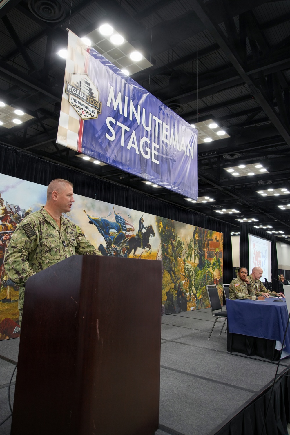 A military officer in Navy camouflage uniform speaks at a podium on the 'Minuteman Stage' at what appears to be the NGAUS (National Guard Association of the United States) 2026 conference in Indianapolis. A large blue banner identifying the stage hangs overhead, while two additional military personnel are seated at a table on the stage behind the speaker. The backdrop features a large panoramic painting depicting historical American military scenes.
