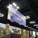 A military officer in Navy camouflage uniform speaks at a podium on the 'Minuteman Stage' at what appears to be the NGAUS (National Guard Association of the United States) 2026 conference in Indianapolis. A large blue banner identifying the stage hangs overhead, while two additional military personnel are seated at a table on the stage behind the speaker. The backdrop features a large panoramic painting depicting historical American military scenes.