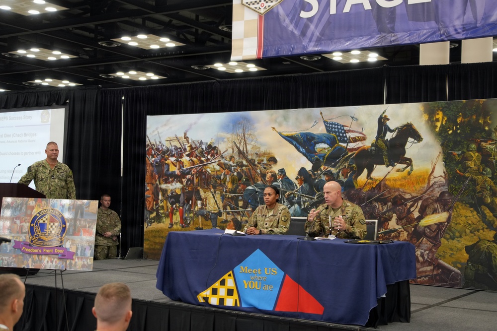 A military panel discussion or briefing is taking place on a stage at what appears to be a conference or convention center. Two soldiers in Army Combat Uniforms are seated at a blue-draped table bearing the slogan 'Meet US where YOU are,' while a Navy officer speaks at a podium to the left. A large mural depicting a Civil War-era battle scene serves as the backdrop.