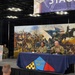 A military panel discussion or briefing is taking place on a stage at what appears to be a conference or convention center. Two soldiers in Army Combat Uniforms are seated at a blue-draped table bearing the slogan 'Meet US where YOU are,' while a Navy officer speaks at a podium to the left. A large mural depicting a Civil War-era battle scene serves as the backdrop.