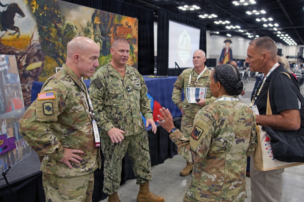 A group of military personnel from multiple branches are engaged in conversation at what appears to be a military convention or expo hall. The group includes U.S. Army soldiers in OCP (Operational Camouflage Pattern) uniforms and a U.S. Navy sailor in NWU (Navy Working Uniform) Type III. They are gathered near a display booth with military-themed artwork and informational materials in a large convention center.