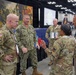 A group of military personnel from multiple branches are engaged in conversation at what appears to be a military convention or expo hall. The group includes U.S. Army soldiers in OCP (Operational Camouflage Pattern) uniforms and a U.S. Navy sailor in NWU (Navy Working Uniform) Type III. They are gathered near a display booth with military-themed artwork and informational materials in a large convention center.