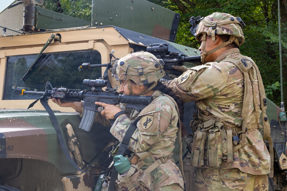 Two soldiers in U.S. Army MultiCam (OCP) uniforms are taking covered positions beside a military HMMWV (Humvee) vehicle. The forward soldier is in a kneeling stance aiming an M4 carbine with optics and accessories, while the second soldier stands behind providing overwatch with a raised weapon. Both wear Advanced Combat Helmets (ACH) with mounted accessories and unit patches visible on their shoulders.