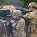 Two soldiers in U.S. Army MultiCam (OCP) uniforms are taking covered positions beside a military HMMWV (Humvee) vehicle. The forward soldier is in a kneeling stance aiming an M4 carbine with optics and accessories, while the second soldier stands behind providing overwatch with a raised weapon. Both wear Advanced Combat Helmets (ACH) with mounted accessories and unit patches visible on their shoulders.
