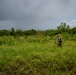 A single individual wearing camouflage clothing and a hat traverses a dense, overgrown grassy field with scattered shrubs and trees in the background. The sky is heavily overcast with dark storm clouds, suggesting an approaching weather system. The scene appears to be a remote, wild landscape, possibly in a tropical or subtropical region.