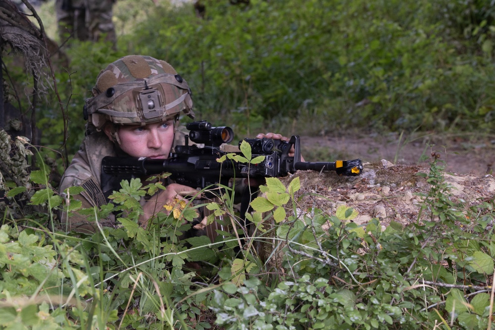 A soldier in full multicam camouflage gear is positioned in a prone or low crouch behind vegetation and a rocky berm, aiming a rifle equipped with optics and accessories. The setting appears to be a wooded or forested training or operational environment. A second figure in camouflage is partially visible in the background.