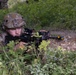 A soldier in full multicam camouflage gear is positioned in a prone or low crouch behind vegetation and a rocky berm, aiming a rifle equipped with optics and accessories. The setting appears to be a wooded or forested training or operational environment. A second figure in camouflage is partially visible in the background.