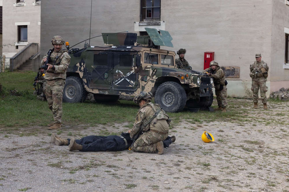 Military personnel in camouflage uniforms are conducting what appears to be a training exercise in an urban environment. One soldier is kneeling over a person lying on the ground, likely performing simulated casualty care or TCCC (Tactical Combat Casualty Care) training. A HMMWV (Humvee) with camouflage paint is positioned nearby while other soldiers maintain security positions.