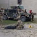 Military personnel in camouflage uniforms are conducting what appears to be a training exercise in an urban environment. One soldier is kneeling over a person lying on the ground, likely performing simulated casualty care or TCCC (Tactical Combat Casualty Care) training. A HMMWV (Humvee) with camouflage paint is positioned nearby while other soldiers maintain security positions.