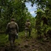 Two military personnel in camouflage uniforms and tactical vests are walking through a dense, green forested area. They are moving away from the camera along what appears to be a trail or clearing between trees. The image captures a patrol or reconnaissance activity in a woodland environment.