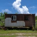 The image depicts what appears to be a military training exercise taking place around a weathered, rust-covered metal container or portable structure. At least two armed personnel in military gear are visible — one taking a prone or crouched firing position at the left corner of the structure, and one or more individuals visible through the right window. The setting is an outdoor training area with overgrown vegetation and a bright, partly cloudy sky.