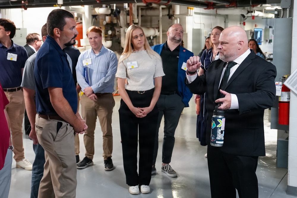 A group of approximately 10 people are gathered in what appears to be an industrial or mechanical facility, such as a utility room or building systems area. A bald man in a black suit is speaking and gesturing to the group, suggesting he is leading a tour or presentation. Most attendees are wearing name badge lanyards, indicating a formal or organized event.