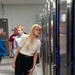 A group of visitors are touring what appears to be a data center or server room facility. A young woman with long blonde hair is prominently in the foreground, leaning in to examine server racks with blue LED lighting. Several other visitors and what appears to be a guide are visible in the background.