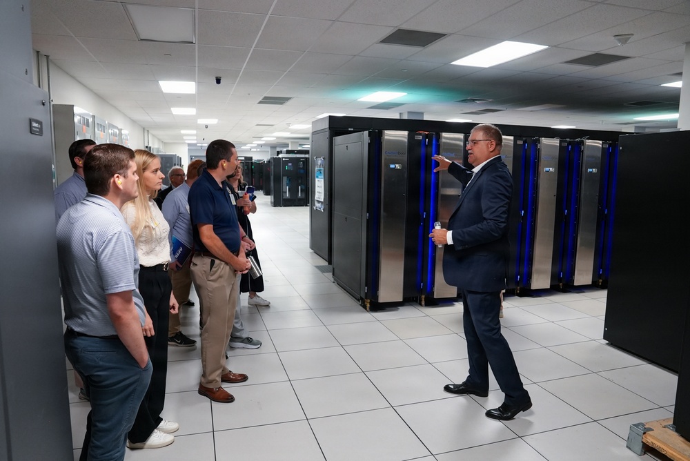 A group of approximately 8-10 people are receiving a guided tour or briefing inside a large data center or server room facility. A man in a dark suit is gesturing toward a row of server racks with distinctive blue LED lighting, appearing to explain or present the equipment. The facility features raised white tile flooring, fluorescent ceiling lighting, and rows of large black server cabinets extending into the background.