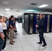 A group of approximately 8-10 people are receiving a guided tour or briefing inside a large data center or server room facility. A man in a dark suit is gesturing toward a row of server racks with distinctive blue LED lighting, appearing to explain or present the equipment. The facility features raised white tile flooring, fluorescent ceiling lighting, and rows of large black server cabinets extending into the background.