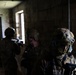 The image shows multiple military personnel in full tactical gear inside a darkened stone or concrete block building. They appear to be conducting a room-clearing or building-entry exercise, with at least one soldier aiming a rifle toward a window. The foreground soldier wears a tactical helmet with mounted equipment and protective eyewear.