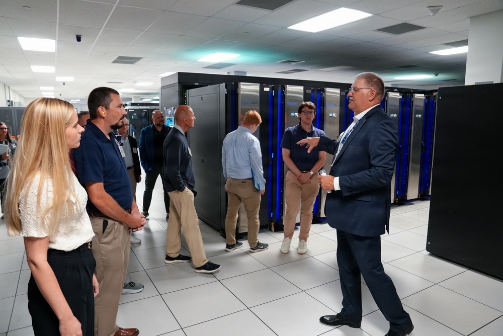 A group of approximately 10 people are receiving a guided tour or briefing inside a large data center or high-performance computing facility. A man in a blue suit, appearing to be the presenter or guide, is speaking to the group while standing near rows of server racks with blue LED lighting. The facility features tall black server cabinets, white tile flooring, and a drop ceiling with fluorescent lighting.