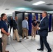 A group of approximately 10 people are receiving a guided tour or briefing inside a large data center or high-performance computing facility. A man in a blue suit, appearing to be the presenter or guide, is speaking to the group while standing near rows of server racks with blue LED lighting. The facility features tall black server cabinets, white tile flooring, and a drop ceiling with fluorescent lighting.