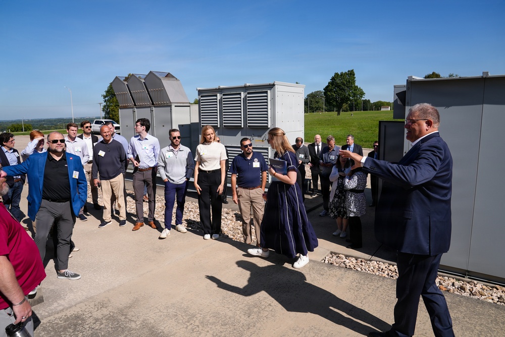 A group of approximately 20 professionals are gathered outdoors at what appears to be an industrial or utility facility for a formal presentation or tour. A man in a navy suit on the right side appears to be addressing the group, while a woman in a dark dress holds materials and stands near the center. The setting includes large industrial equipment units, likely generators or HVAC systems, on a concrete pad surrounded by gravel and green landscape.