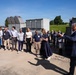 A group of approximately 20 professionals are gathered outdoors at what appears to be an industrial or utility facility for a formal presentation or tour. A man in a navy suit on the right side appears to be addressing the group, while a woman in a dark dress holds materials and stands near the center. The setting includes large industrial equipment units, likely generators or HVAC systems, on a concrete pad surrounded by gravel and green landscape.