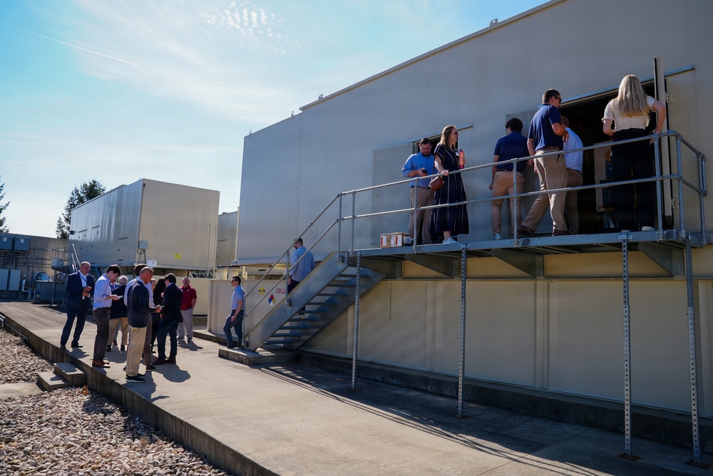 A group of people are conducting what appears to be a facility tour or inspection at an industrial or utility building. Some individuals are gathered on an elevated metal platform/walkway at the entrance of the structure, while others remain on the ground level in a separate discussion group. The setting appears to be an outdoor industrial compound on a clear, sunny day.