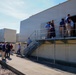 A group of people are conducting what appears to be a facility tour or inspection at an industrial or utility building. Some individuals are gathered on an elevated metal platform/walkway at the entrance of the structure, while others remain on the ground level in a separate discussion group. The setting appears to be an outdoor industrial compound on a clear, sunny day.