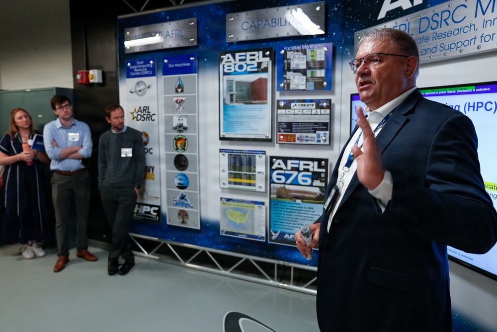 A man in a dark suit is presenting or speaking at what appears to be an AFRL (Air Force Research Laboratory) DSRC (DoD Supercomputing Resource Center) briefing or tour. He stands in front of a large display board featuring logos, facility information, and references to HPC (High Performance Computing) systems including AFRL 802 and AFRL 676. Three attendees with name badges observe from the background.