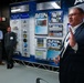A man in a dark suit is presenting or speaking at what appears to be an AFRL (Air Force Research Laboratory) DSRC (DoD Supercomputing Resource Center) briefing or tour. He stands in front of a large display board featuring logos, facility information, and references to HPC (High Performance Computing) systems including AFRL 802 and AFRL 676. Three attendees with name badges observe from the background.