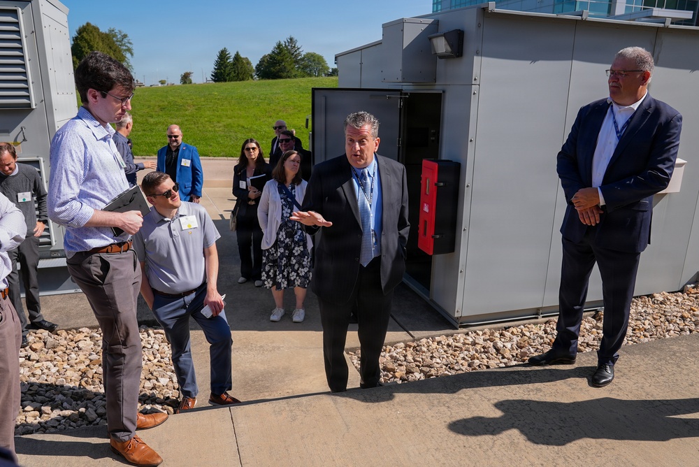 A group of approximately 10-12 professionals in business attire are gathered outdoors at what appears to be a facility tour or site visit. A central figure in a gray suit and blue tie is actively speaking and gesturing, suggesting he is leading the tour or presentation. The setting includes industrial or utility equipment enclosures, gravel landscaping, and a manicured green lawn in the background.